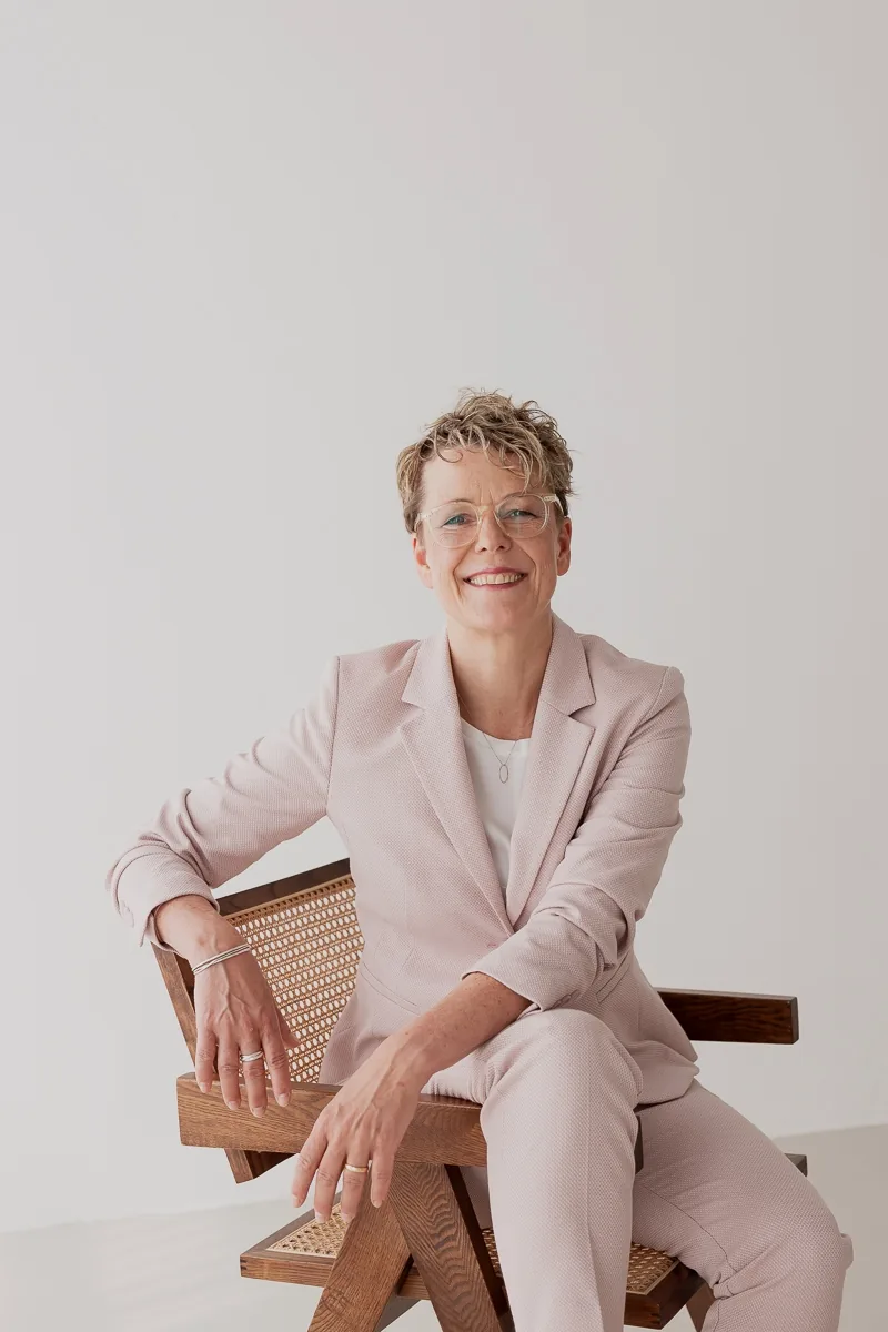 Britta Wein sitzt lässig im rosafarbenen Anzug auf einem Holzstuhl und blickt in die Kamera.